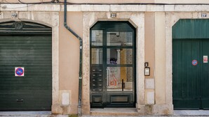 A building with a closed green door, a closed shutter door, and a numbered address sign. - São Mamede Downtown Apartment (Lisbon)