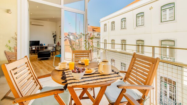 A balcony with wooden chairs and a table set with breakfast items, overlooking a cityscape.