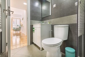 A modern bathroom with a white toilet, a cabinet with a green emergency sign, and a teal trash bin. - Amazing Castle Views in Historic Center (Lisbon)
