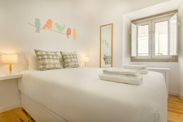A bedroom with a large bed, two bedside tables with lamps, a window with curtains, and a wall decoration of birds. - Amazing Castle Views in Historic Center (Lisbon)