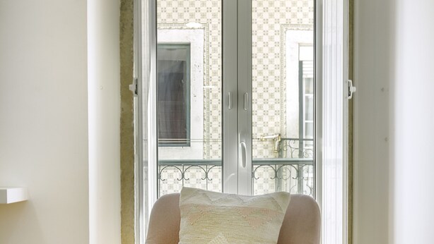 A room with a wooden floor, a chair with a cushion, a small table with a book, and a door leading to a balcony with patterned tiles. - Amazing Castle Views in Historic Center (Lisbon)