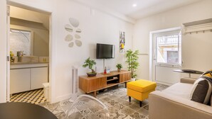 A modern living room with a sofa, a TV, a small table, and a yellow ottoman. There is a kitchen area with a white countertop and a tiled backsplash. - Steps Lisbon @ Historical Center (Lisbon)