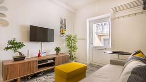 A modern living room with a flat-screen TV, wooden entertainment unit, yellow ottoman, and a window with a view of a building. - Steps Lisbon @ Historical Center (Lisbon)