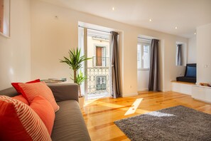 Sala de estar moderna com sofá cinzento, almofadas vermelhas, planta em vaso e soalho de madeira.