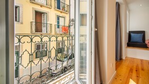 A room with a large window, a wooden floor, a bed with a blue pillow, and a balcony with a metal railing. - Bica Historical Tram Apartment (Lisbon)