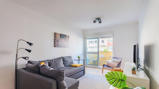 Ein modernes Wohnzimmer mit grauem Sofa, einer Stehlampe, einem kleinen Tisch mit Pflanze und einem Fernseher auf einem Schrank.