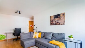 Ein modernes Wohnzimmer mit einem grauen Ecksofa, einem Glastisch, einem schwarzen Bürostuhl, einem kleinen Topfpflanzen und einem Bild an der Wand.
