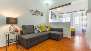 A modern living room with a grey sectional sofa, a glass coffee table, and a lamp. There are framed pictures on the wall and a window with curtains. - Paraíso Alfama (Lisbon)