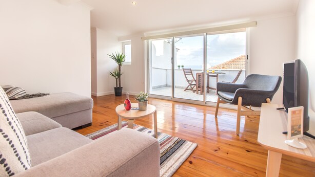 Una sala moderna con un sofá gris, una mesita redonda y un televisor. Hay un balcón con una mesa y sillas, y una planta en maceta.