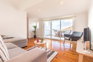 Una sala moderna con un sofá gris, una mesita redonda y un televisor. Hay un balcón con una mesa y sillas, y una planta en maceta.
