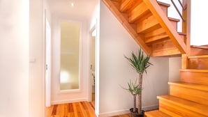 Una escalera de madera con diseño moderno, una planta en maceta y un pasillo con suelo de madera y paredes blancas.