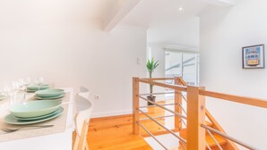 Un interior moderno con una escalera de madera, un comedor con platos verdes y sillas blancas, y una planta en maceta.