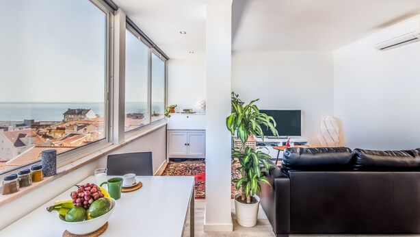 A modern living room with a large window, a black leather sofa, a dining table with fruits and a bowl, and a potted plant. - Graça Amazing River Views (Lisbon)