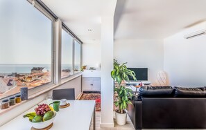 A modern living room with a large window, a black leather sofa, a dining table with fruits and a bowl, and a potted plant. - Graça Amazing River Views (Lisbon)