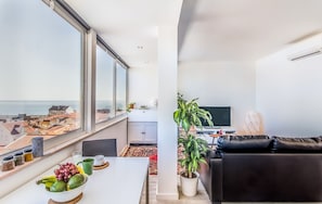 A modern apartment with a balcony, a dining area with a table and chairs, a black leather sofa, a potted plant, and a television.