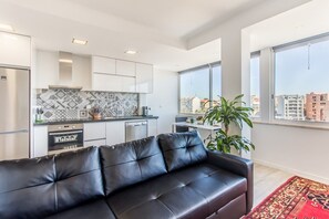 A modern apartment with a kitchen, a black leather sofa, a red rug, and large windows with a city view.