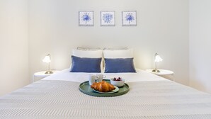 Un letto rifatto con cura, con un vassoio pieno di croissant, tazze di caffè e frutti di bosco, affiancato da due comodini con lampade, in una stanza dal design semplice e pulito.