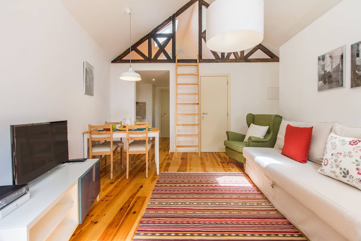 Una sala de estar moderna con piso de madera, un sofá blanco, un sillón verde, un televisor, una mesa de comedor con sillas y una escalera que da a un altillo.