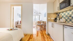 Una camera d'albergo compatta con pavimento in legno, un letto con copripiumino bianco, una zona pranzo con tavolo e sedie e un angolo cottura con forno a microonde e macchina da caffè.