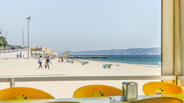 A beach view from a window with yellow chairs and a table. - Luxury Beach Experience in Lisbon (Oeiras)