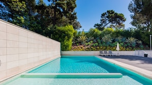 A rectangular swimming pool with clear blue water, surrounded by a tiled wall and a landscaped area with various plants and trees. - Luxury Beach Experience in Lisbon (Oeiras)