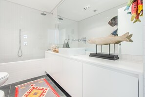 A modern bathroom with a glass shower enclosure, a white sink with a fish sculpture, and a patterned rug. - Luxury Beach Experience in Lisbon (Oeiras)