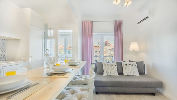 A modern living room with a grey sofa, a dining table set with plates and glasses, and a view of buildings through large windows. - Roma Cozy Flat (Lisbon)