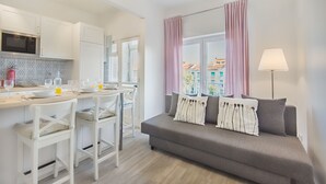 A modern kitchen with white cabinets and a dining area with a table and chairs. There is a grey sofa with decorative pillows and a window with pink curtains. - Roma Cozy Flat (Lisbon)