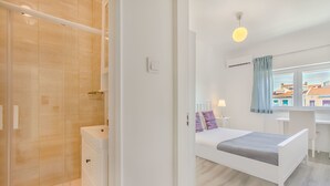 A modern bedroom with a shower, a bed, a desk, and a view of buildings outside. - Roma Cozy Flat (Lisbon)