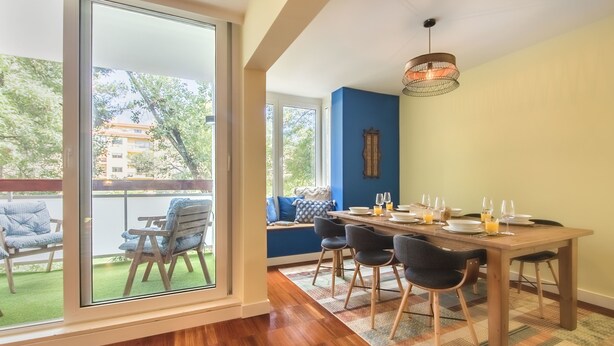 A dining area with a wooden table set for four, a blue accent wall, and a view of the outdoors through large sliding glass doors. - 347 Acácias Flat (Cascais)