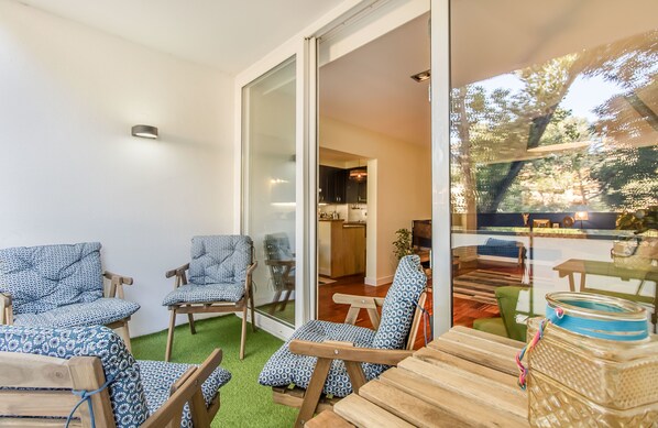 A modern outdoor seating area with patterned chairs and a wooden table, a glass door leading to a kitchen, and a view of a garden through the glass. - 347 Acácias Flat (Cascais)
