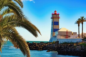 A lighthouse with a red top, situated next to a rocky shoreline with palm trees. - 347 Acácias Flat (Cascais)