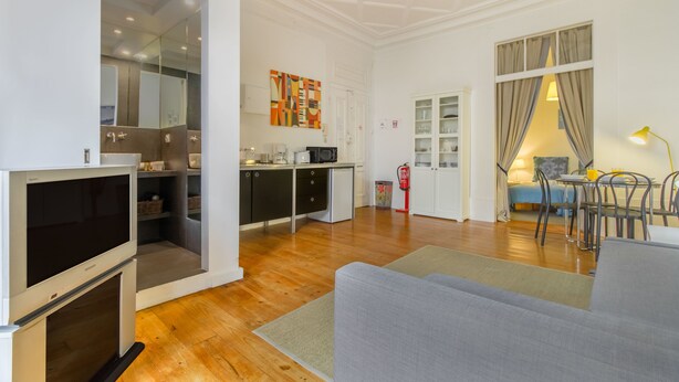 Un salon moderne avec un canapé gris, un plancher en bois, un téléviseur à écran plat, un coin cuisine doté de placards et d’un four à micro-ondes, et un coin repas avec une table et des chaises.