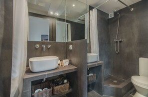 A modern bathroom with a large mirror, a white sink, a shower, and a toilet. There are wicker baskets under the sink and a towel rack above it.