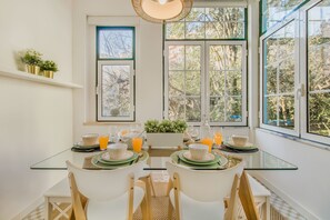 A breakfast table set with green plates, glasses, and orange juice, with a view of trees through large windows. - Graça Trendy Flat (Lisbon)