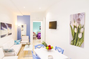Un salón luminoso con un sofá blanco, una mesa de comedor con un frutero y un cuadro grande de tulipanes.