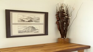 Una obra de arte en blanco y negro con paisaje en un marco, un estante de madera debajo y un jarrón con ramas secas.