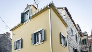 Un edificio amarillo con tres ventanas y contraventanas, una antena parabólica y un muro de piedra.