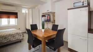 A compact room with a wooden dining table, black chairs, a bed with patterned bedding, a small potted plant, a mounted TV, and a microwave on a cabinet. - Nera Luxury Apartments (Split)