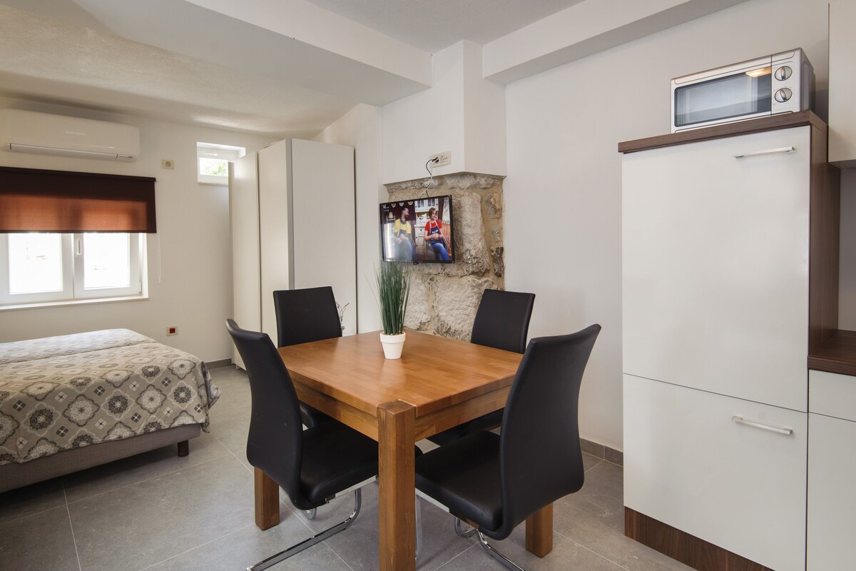 Une pièce de taille modeste, avec une table à manger en bois, des chaises noires, un lit avec une literie à motifs, une petite plante en pot, un téléviseur fixé au mur et un four à micro-ondes posé sur un meuble.