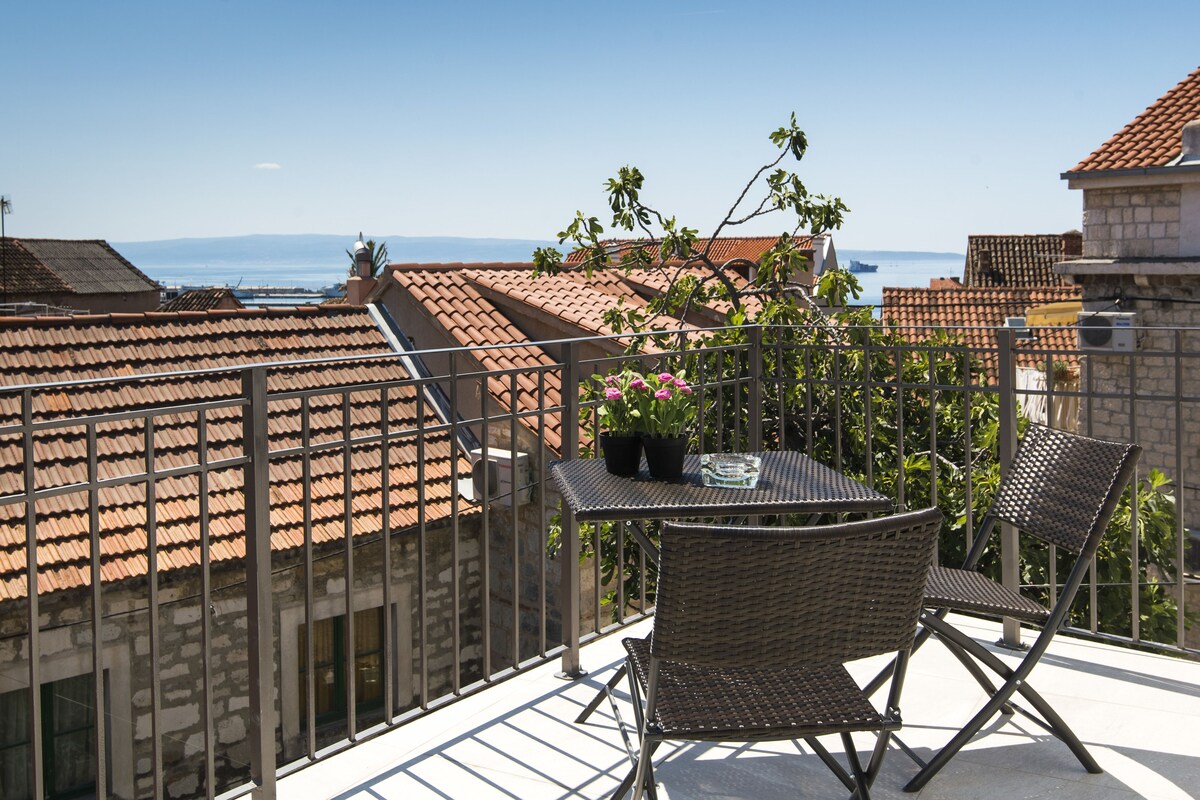 Un balcon avec une table et des chaises, donnant sur un paysage urbain aux toits de tuiles et offrant une vue sur la mer.
