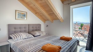 A bedroom with a large bed, a headboard, a window with a view of buildings, and a balcony with a table and chairs. - Nera Luxury Apartments (Split)