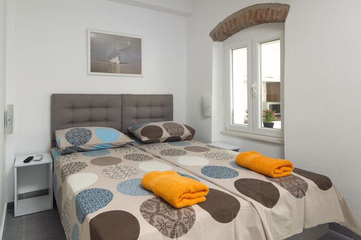 Un lit bien fait, recouvert d’une couvre-lit à motifs, une table de chevet avec une télécommande, et une fenêtre donnant sur l’extérieur.