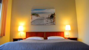 Ein Hotelzimmer mit einem Bett, zwei Nachttischlampen, einem Holzkopfteil und einem Wandbild mit einer Strandansicht.