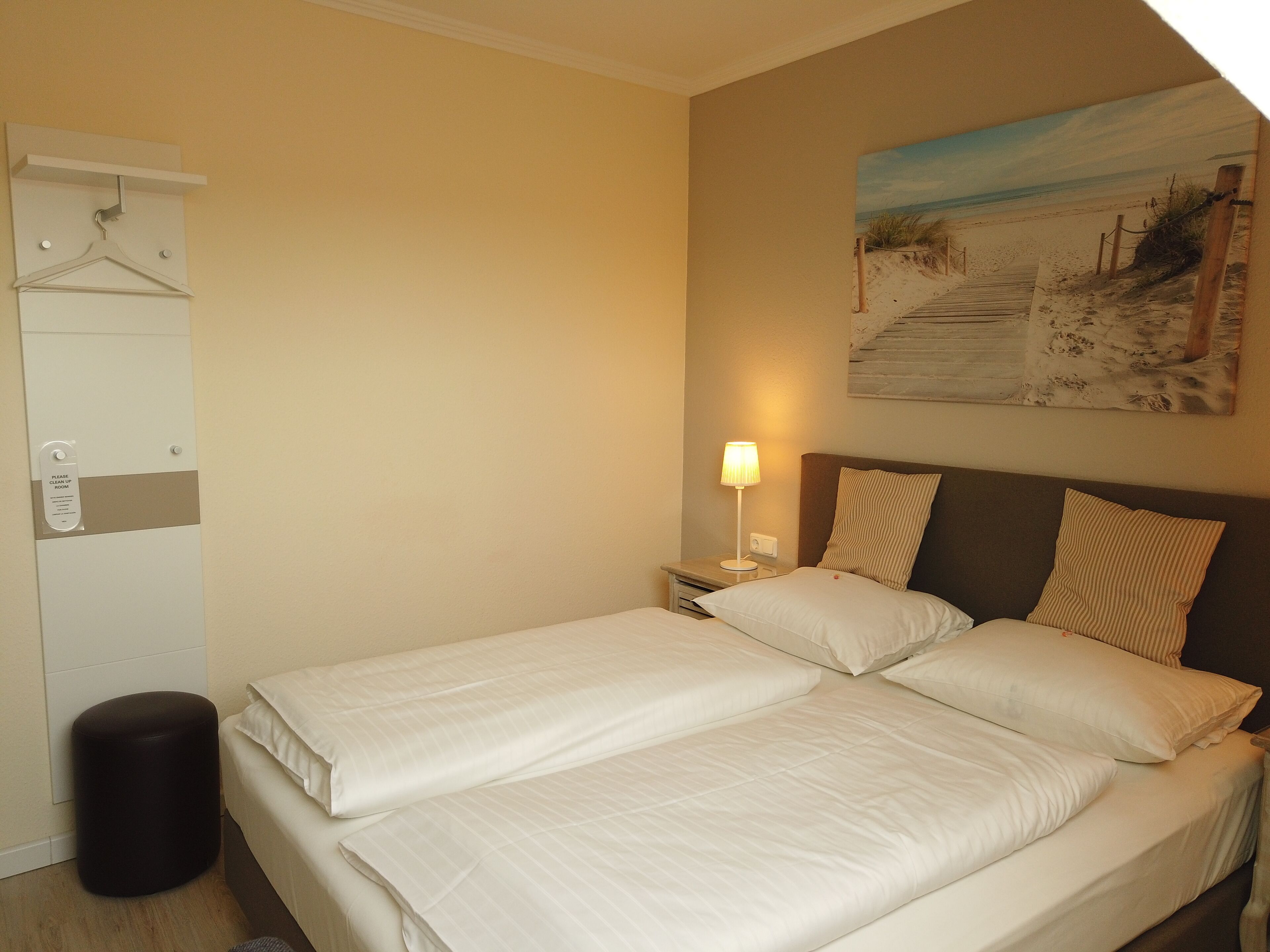 Ein Hotelzimmer mit einem Bett, einem Nachttisch, einer Lampe, einem Strandbild und einem Kleiderhaken.