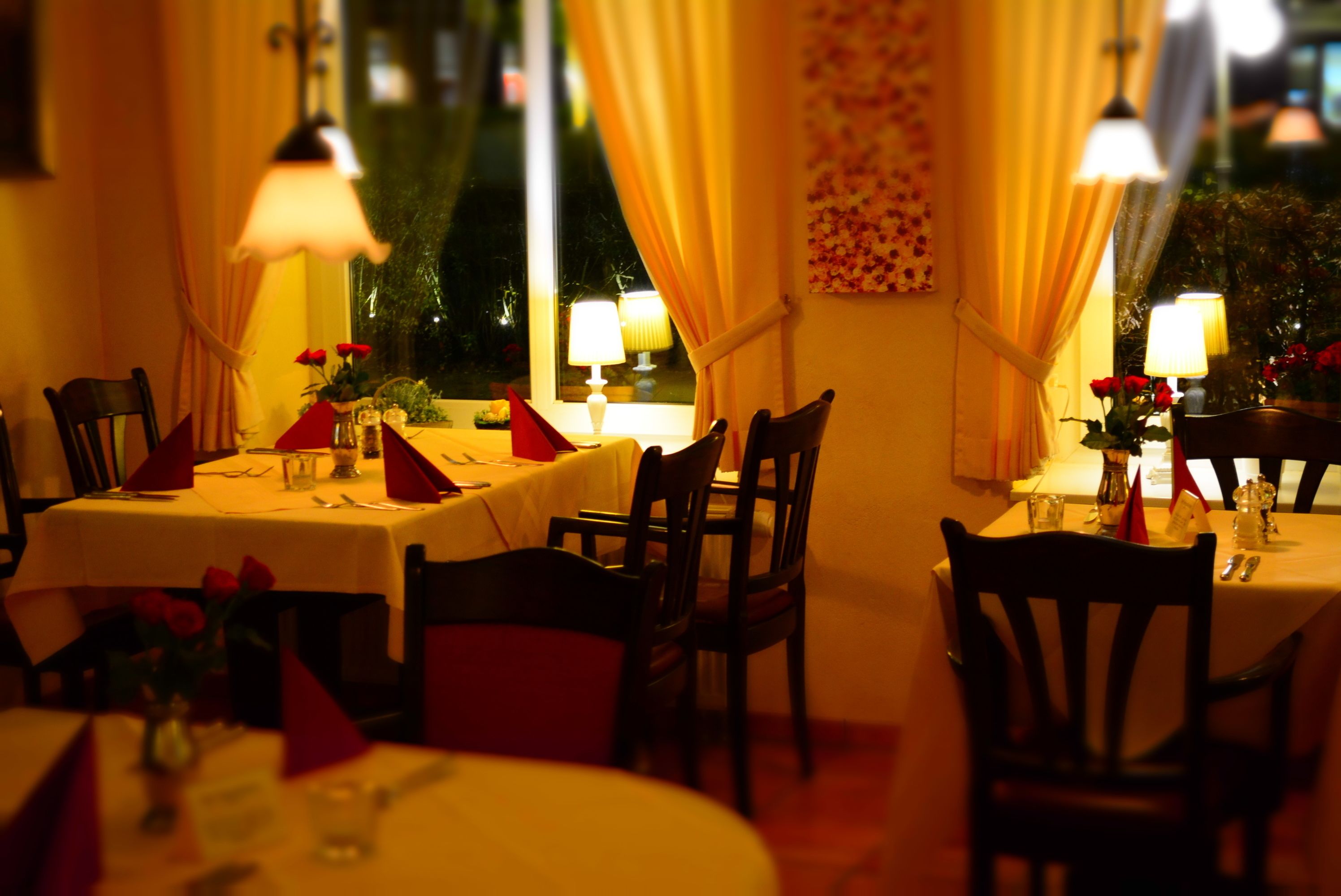 A dining area with tables set for two, chairs, and table lamps.