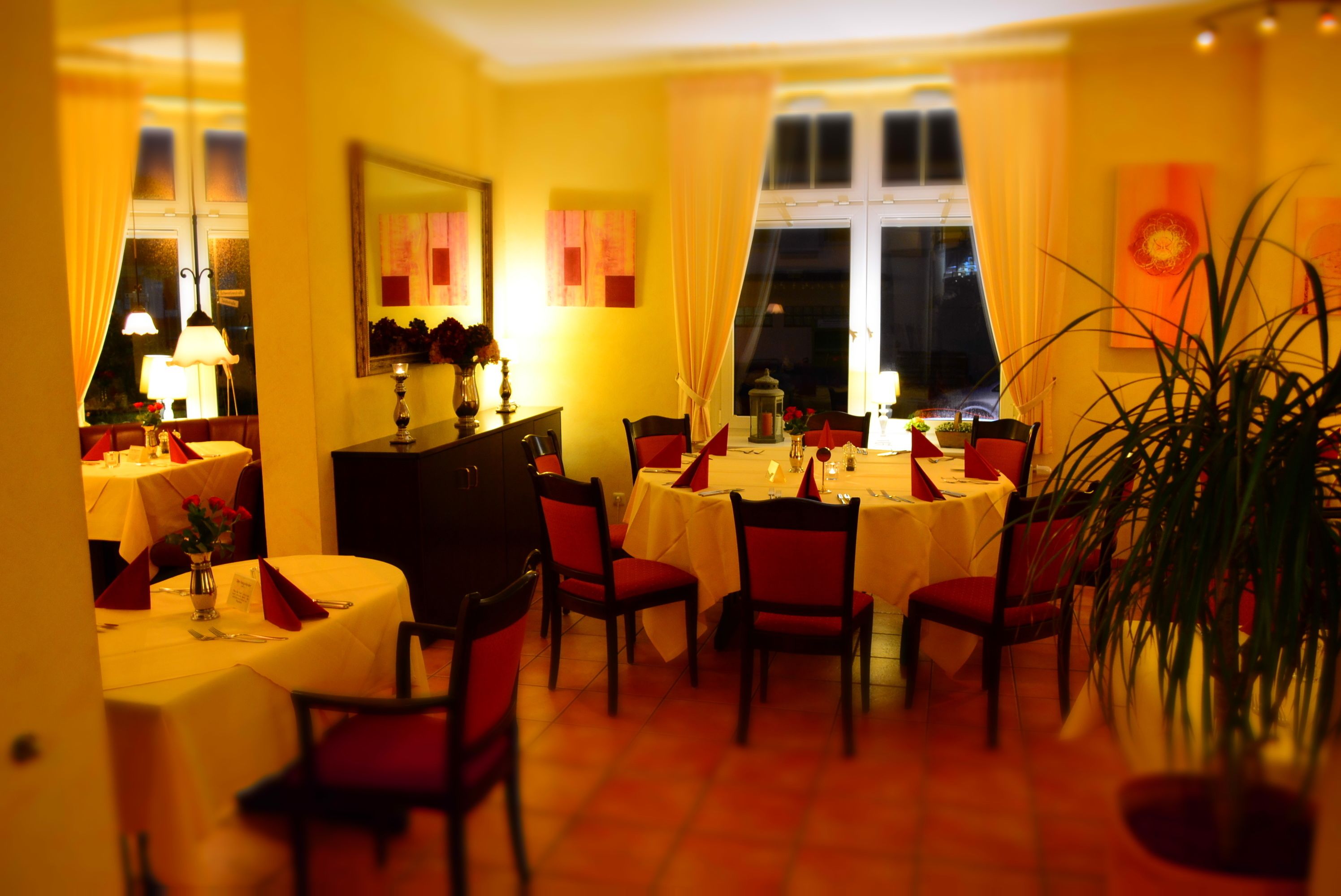 A dining area with tables set for four, chairs, a vase with flowers, and a large window with curtains.
