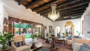 A spacious living room with a high ceiling, wooden beams, and a chandelier. There are several pieces of furniture, including a sofa, armchairs, and a coffee table. The room has large windows and doors leading to an outdoor area with plants.