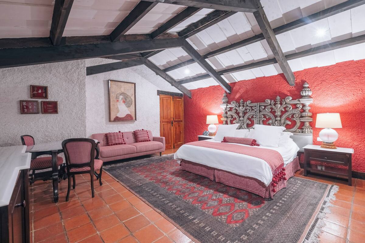 Un dormitorio con una cama grande, un escritorio y una silla. La pared es roja y el techo, blanco.