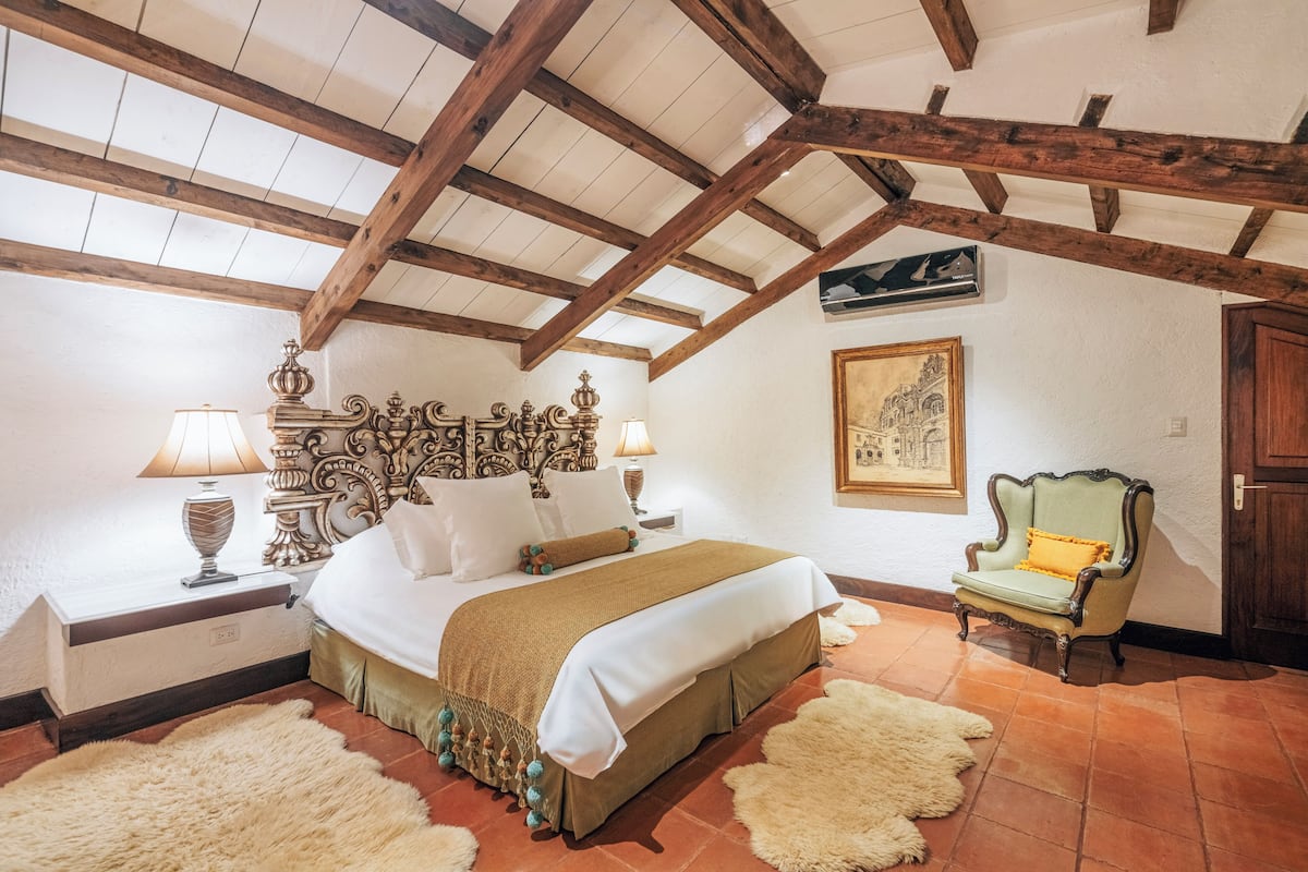Un dormitorio con techo de madera, una cama con edredón blanco y marrón, dos lámparas de noche, una silla y un cuadro en la pared.